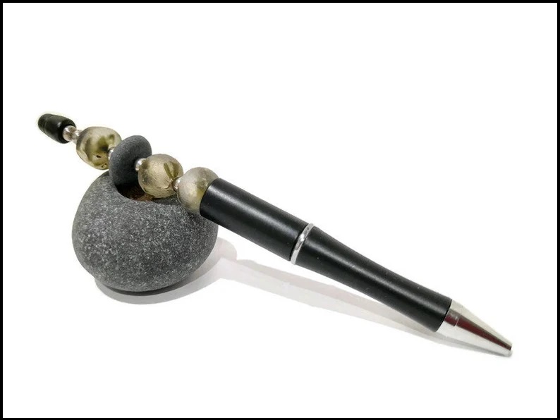 Beach stone and recycled glass beads make up the top part of a pen. The pen is leaning against a small grey stone.
