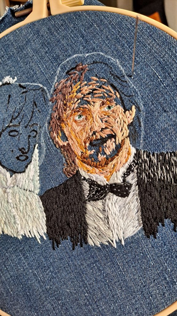 A work of embroidery in mid-production inside an embroidery hoop. The fabric is reused denim and the artist is stitching a couple's wedding portrait