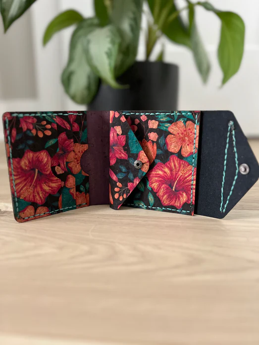 A handmade cork wallet with printed flowers on it sits on top of a wooden table with a plant in the background.