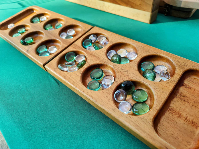 A handmade wooden mancala board sits open on a teal-green table, full of glass playing pieces.

