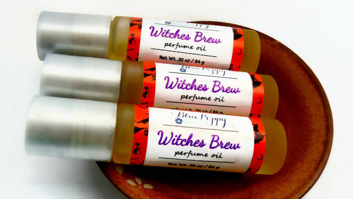 A picture of three perfume rollers in a small wooden dish. Each roller contains a perfume that smells like patchouli, cinnamon, and cedarwood. The roller bottles have orange, white, and purple labels on them. The labels read "Witches Brew Perfume Oil" and features the shop name in a pretty cursive font. 
