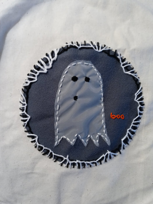 A close up of an embroidered patch on a cotton tote bag. The patch shows a small ghost, carefully stitched with it's mouth in a "boo" shape. In small orange letters, the spirit says "boo" in lowercase. He's trying his best. His best is pretty good.