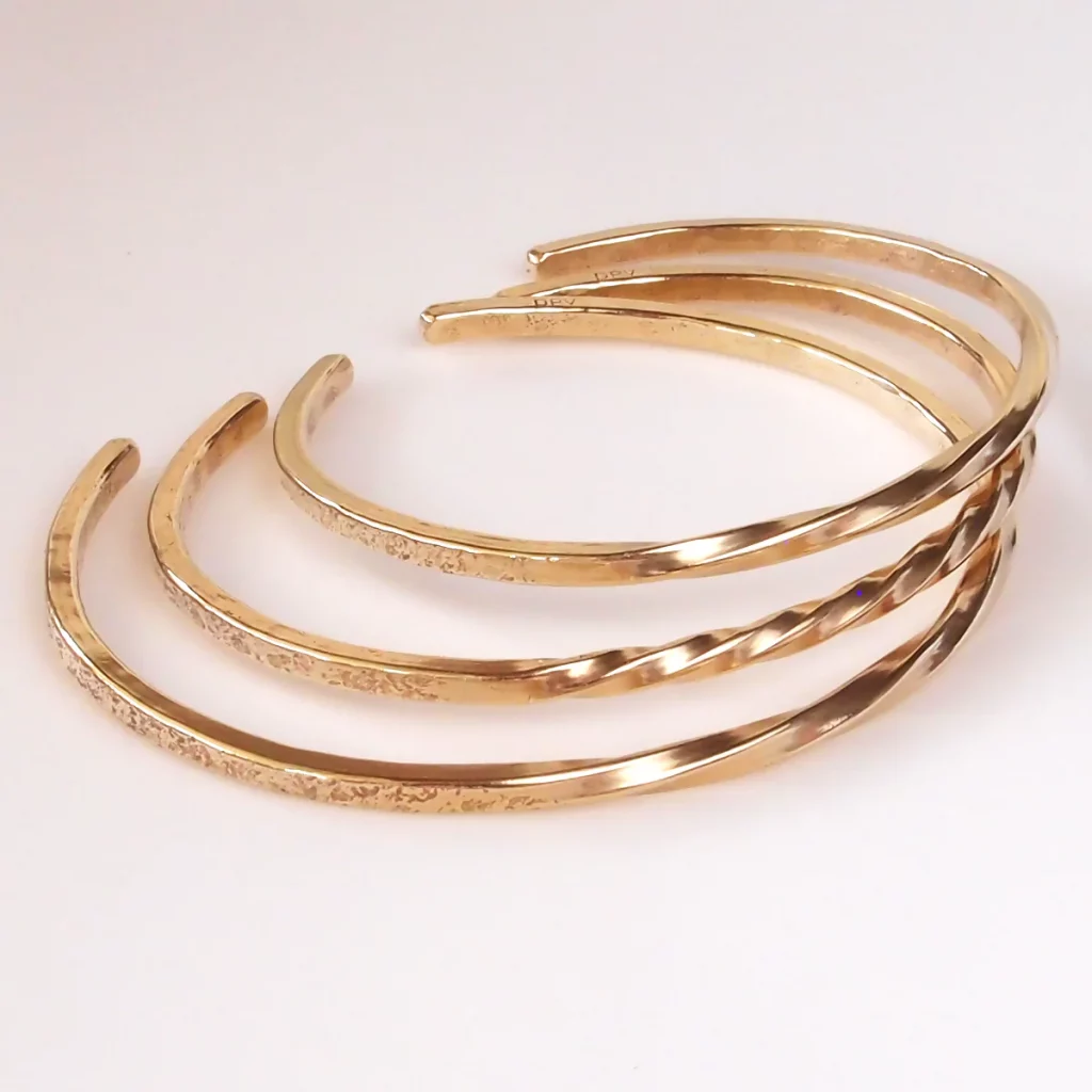 Shows a stack of 3 shiny bronze bracelets in different sizes. They are unadorned and minimalist and not too thick with small twists on the top of the wrist and an open cuff