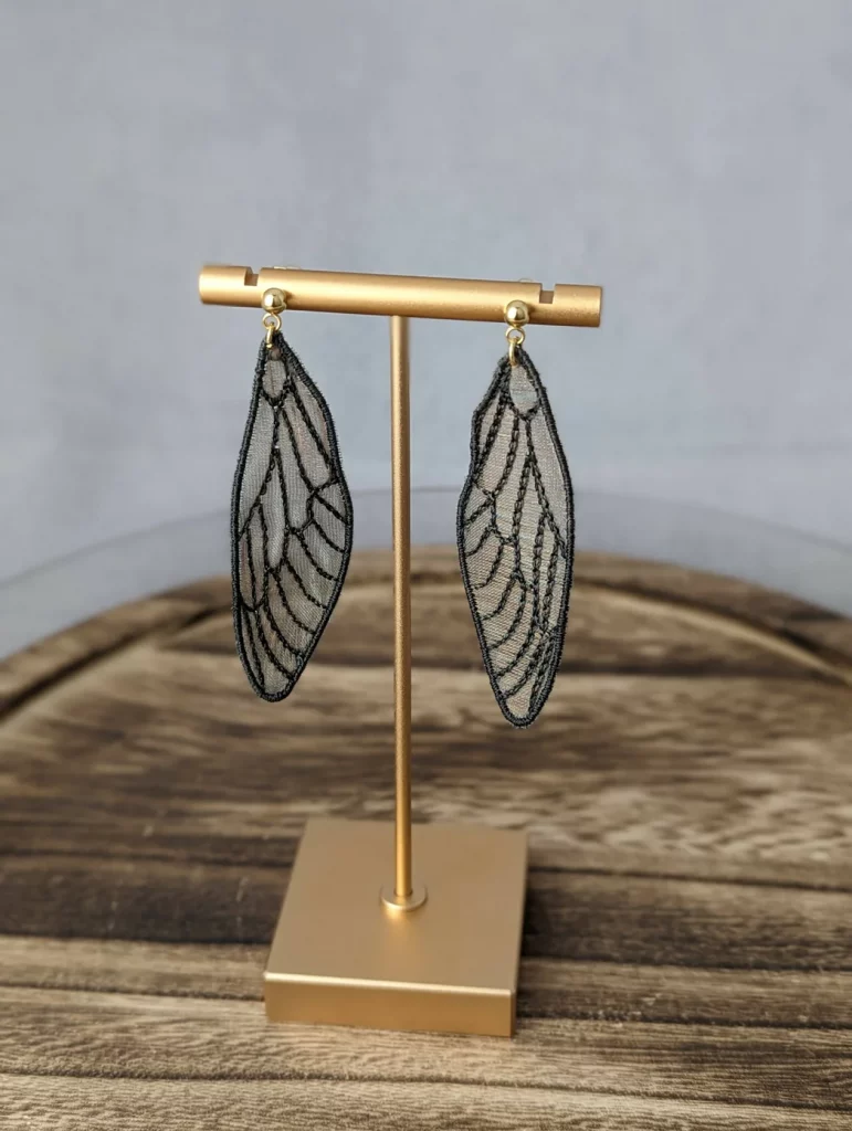 Lacy cicada wings in a natural translucent material with black outlining as dangly earrings hanging from a brass display stand