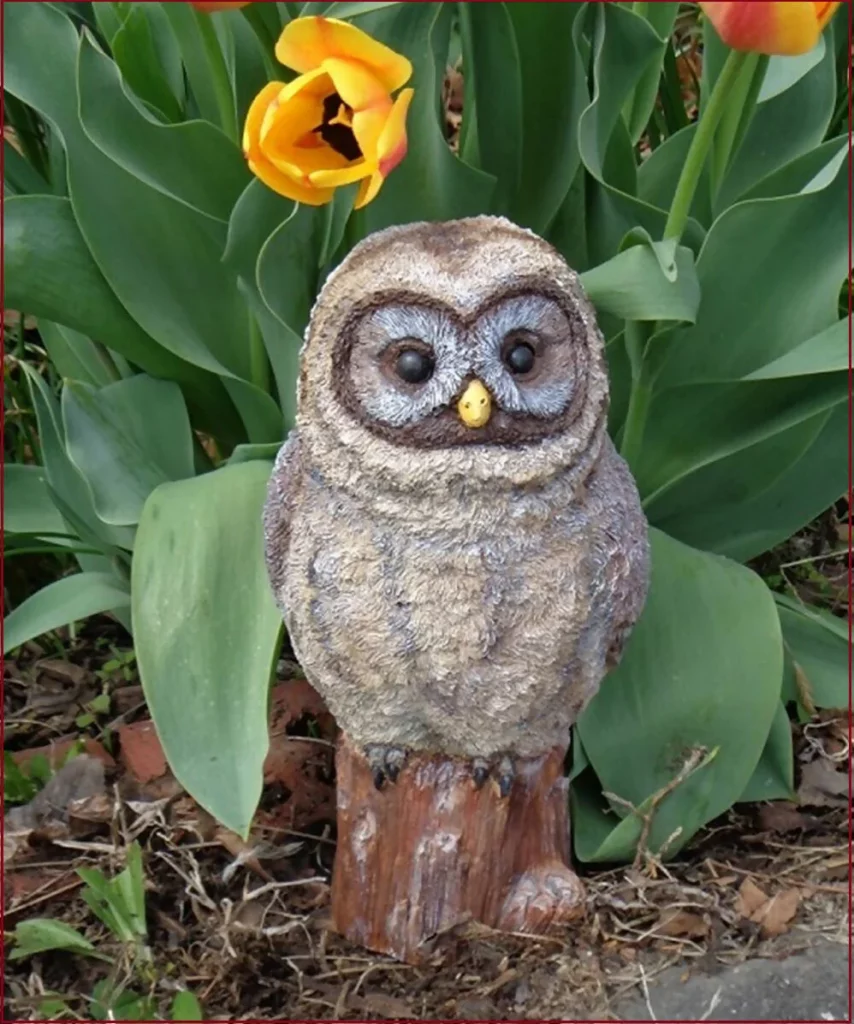 A concrete full color owl statue in a garden in front of a bed of tulips. The owl looks very friendly but realistic