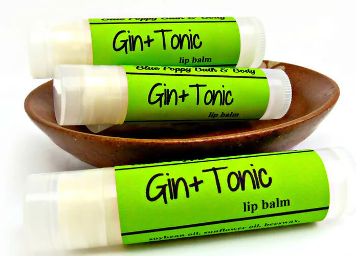 A small wooden bowl on a white background is overflowing with lime green lip balm containers that read "Gin + Tonic"