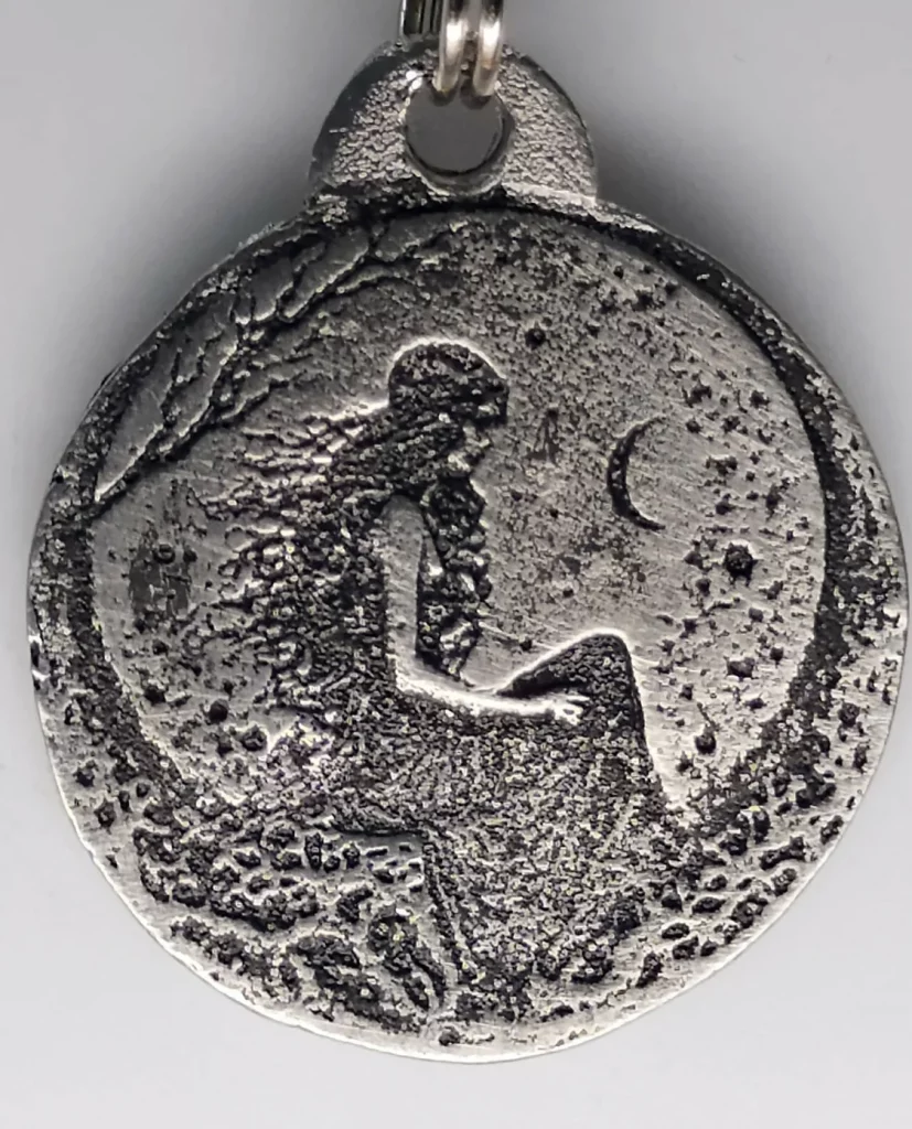 A round, rough-cast pewter pendant of a maiden goddess sitting in profile and a crescent moon