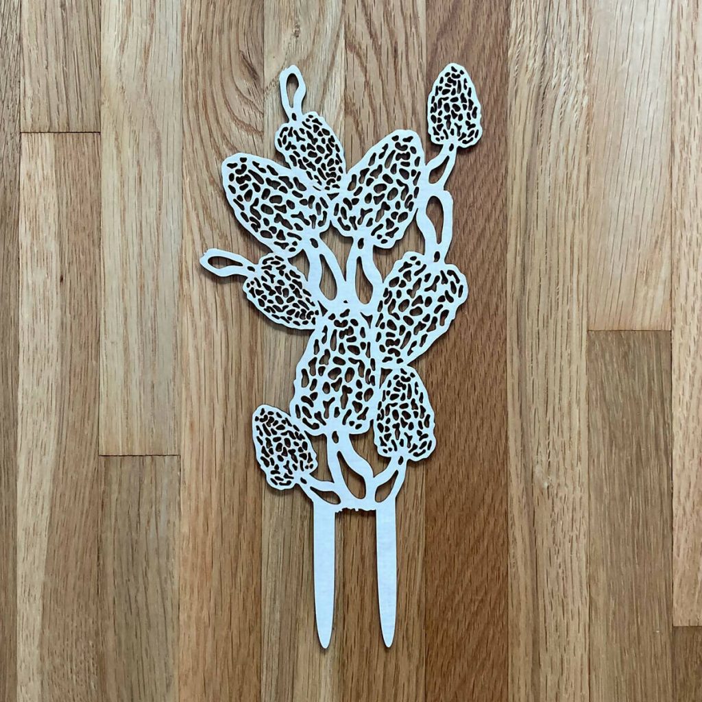 A plant trellis made from intricately laser-cut thin wood in a light color. The decoration shows lacy morel mushrooms