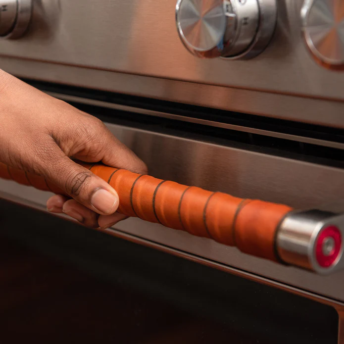 The handle of a stainless steal oven has been decorated with a strip of leather wound around it. A hand is opening the oven.