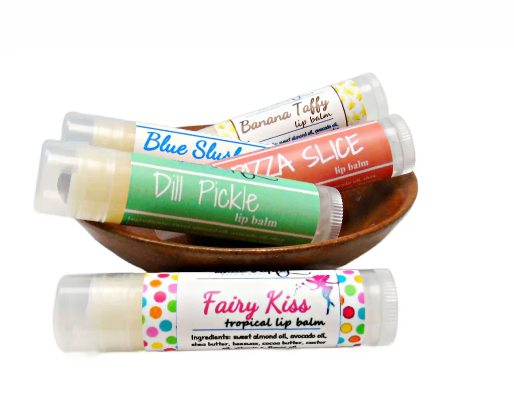 A wooden bowl with four sticks of lip balm in it. Each have bright labels and say "Banana Taffy," "Blue Slush," "Pizza Slice," and "Dill Pickle." There's one on the surface in front of the bowl that says "Fairy Kiss."