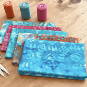 5 patterned fabrics laying folded on top of each other on a light wood table. The fabrics vary in shades of light blue, orange and blue, and pink. The patterns look like watercolor splotches. Behind the fabric are three spools of orange, light blue, and pink thread. 