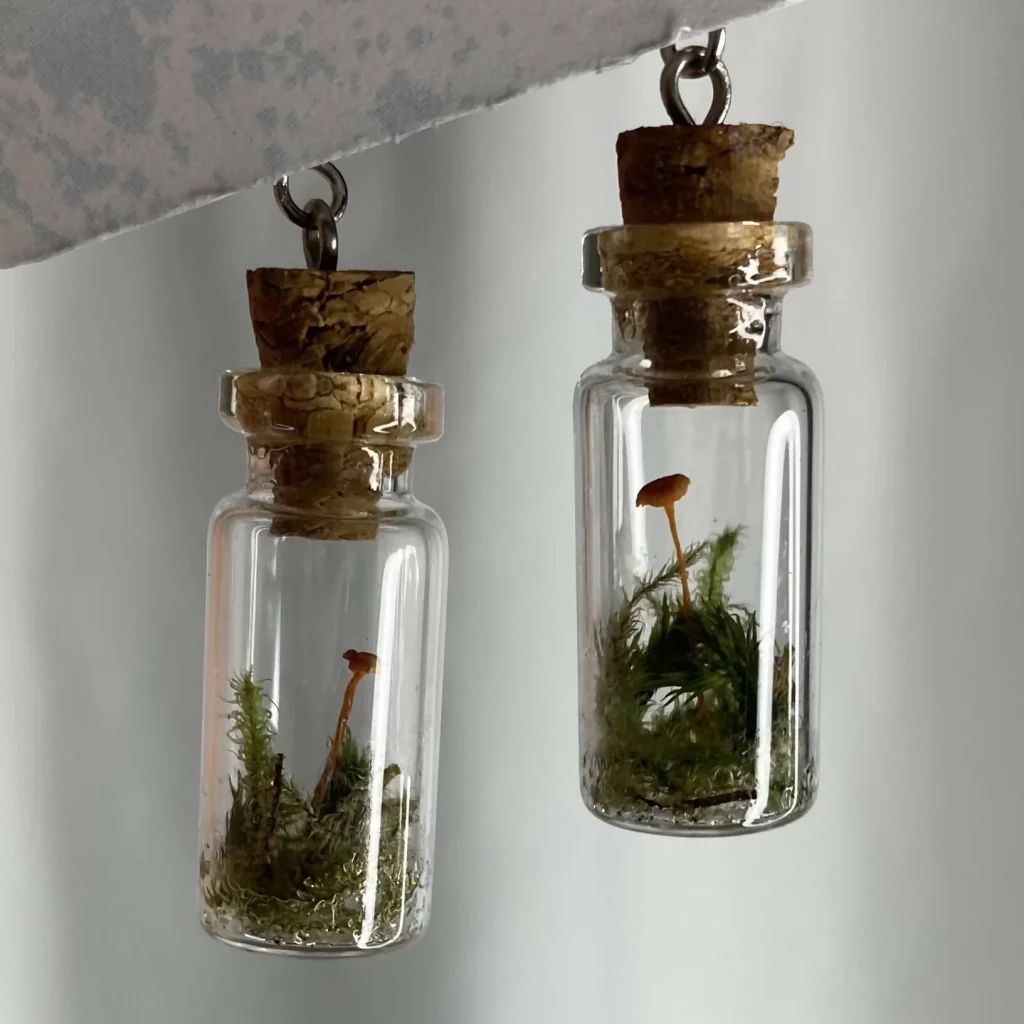 Two tiny corked bottles hang from earring loops. They contain tiny bits of moss and very tiny mushrooms.
