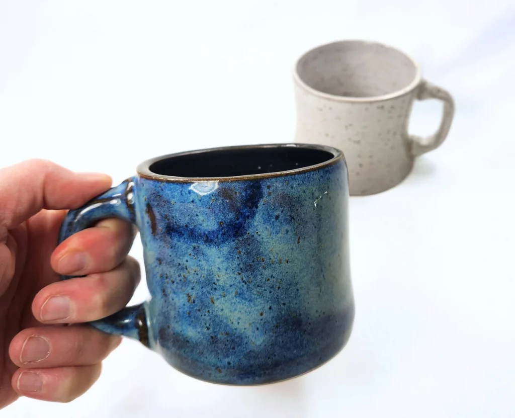 A hand from the left holds a short blue mug. Behind it, on a surface, is a similar mug but in white.