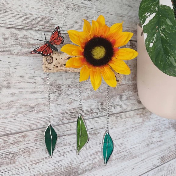 A piece of driftwood hangs from a wooden wall, decorated with a sunflower, a monarch butterfly, and three hanging stained glass leaves.