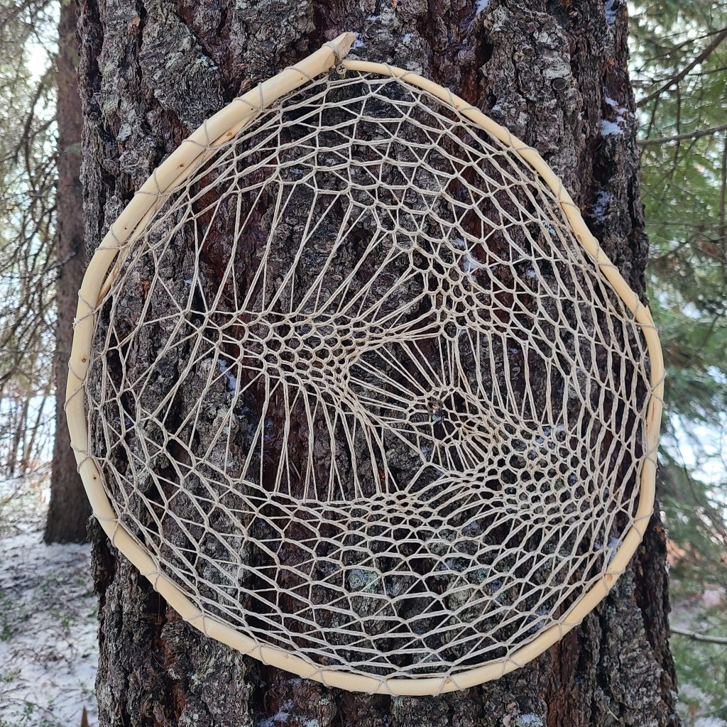 Woven sculpture dream catcher by Brent Joly. Circular cedar and hemp weaving against trunk of tree. 