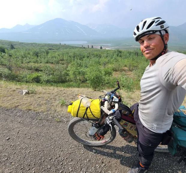 Artisan and woven sculptor, Brent Joly, on his cycle on a lonely Canadian road with a lake and mountains nearby.