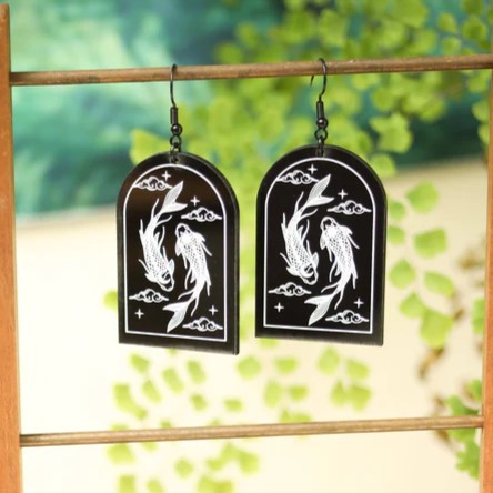 Two arch shaped earrings hanging from a wooden dowel stand. They are black with a clear white design of two koi fish swimming around each other with clouds around them.
