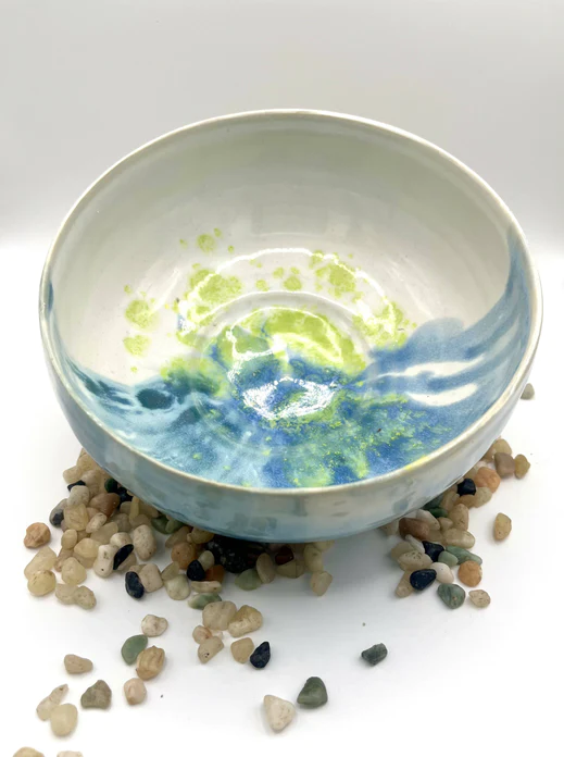 A white ceramic bowl with blue, swirly glaze covering half of it, sits on a white table with some small sand color pebbles around it.