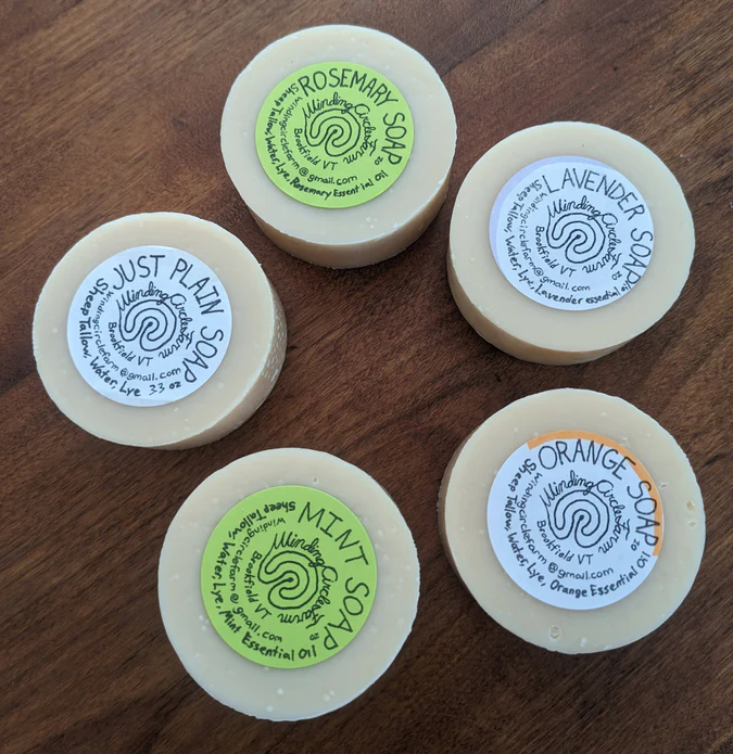 Five round soap bars sitting in a circle on a brown table. Each labeled with a small round sticker, Rosemary, Lavender, Orange, Mint, and Just Plain Soap.
