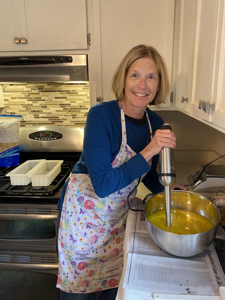 Sheri from Blue Poppy Bath and Body Works mixing a small batch of yellow lemongrass soap with a stick blender in a stainless steel bowl in her clean and organized kitchen. 