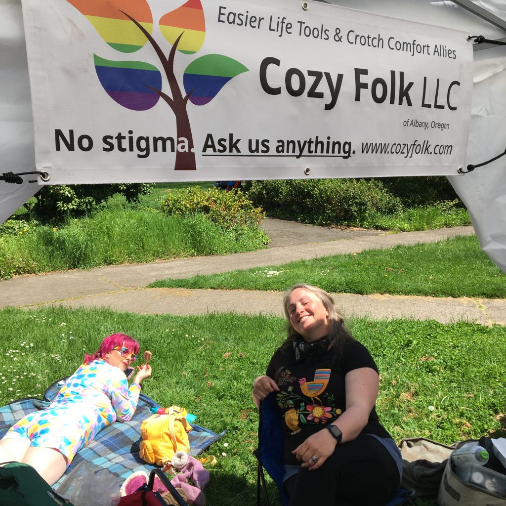 The artisans pose outdoors with their company banner above that says "Cozy Folk LLC" with "No stigma. Ask us anything" below.