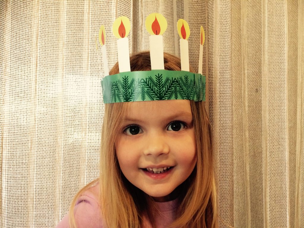A young blonde girl wearing a paper cut out printed St. Lucia crown, with a green garland headband and yellow and white colored candles. 