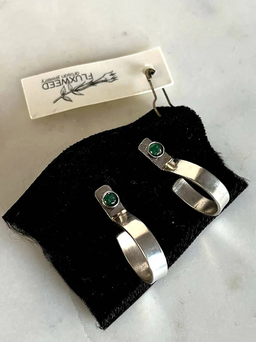 On top of a small black velvet bag labeled Fluxweed Artisan Jewelry, sit two sterling silver loop-like earrings with emerald studs.
