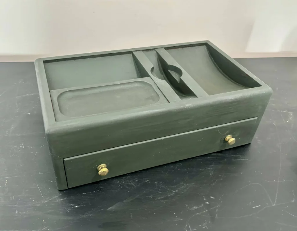 A sage green vintage jewelry box on a table. The top has sections specially for random things - dish for coins, space for watch, etc. There's a drawer at the bottom with two gold drawer pulls.