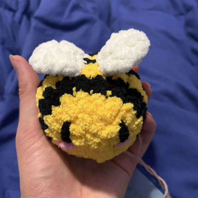 A hand holding a yellow and black crochet bumble bee over a purple blanket background.
