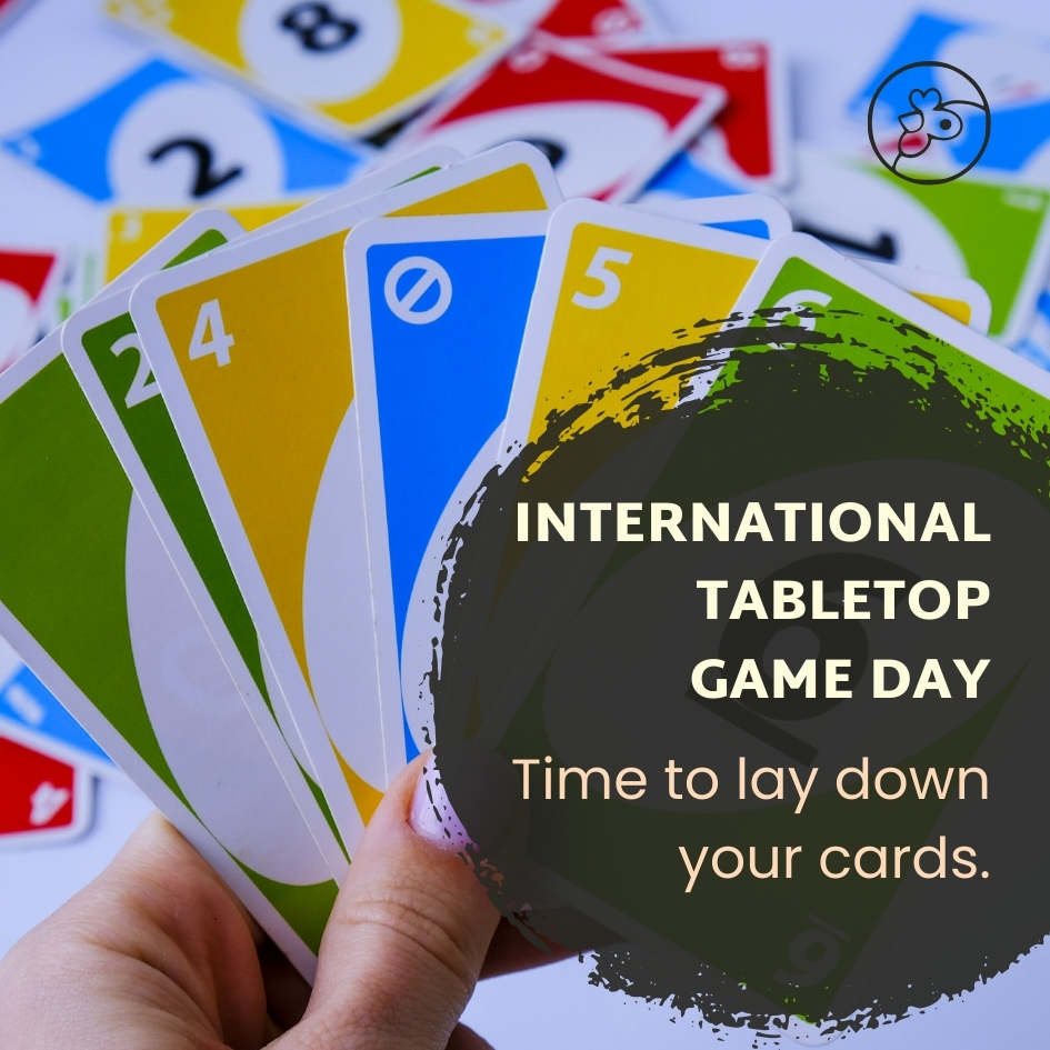 Colorful Uno cards in a person's hand with more cards on the table with a dark circle and white text that reads International Tabletops Games Day Time To Lay Down Your Cards.