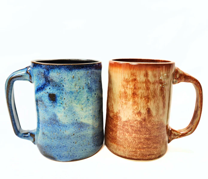 Two handmade ceramic mugs. The left has a blue glaze and the right has an orange-copper glaze on it.