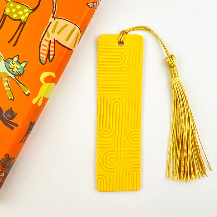 A bright yellow bookmark next to an orange book with yellow cat illustrations on it. The bookmark has wavy arched impressions in it and a yellow tassle.