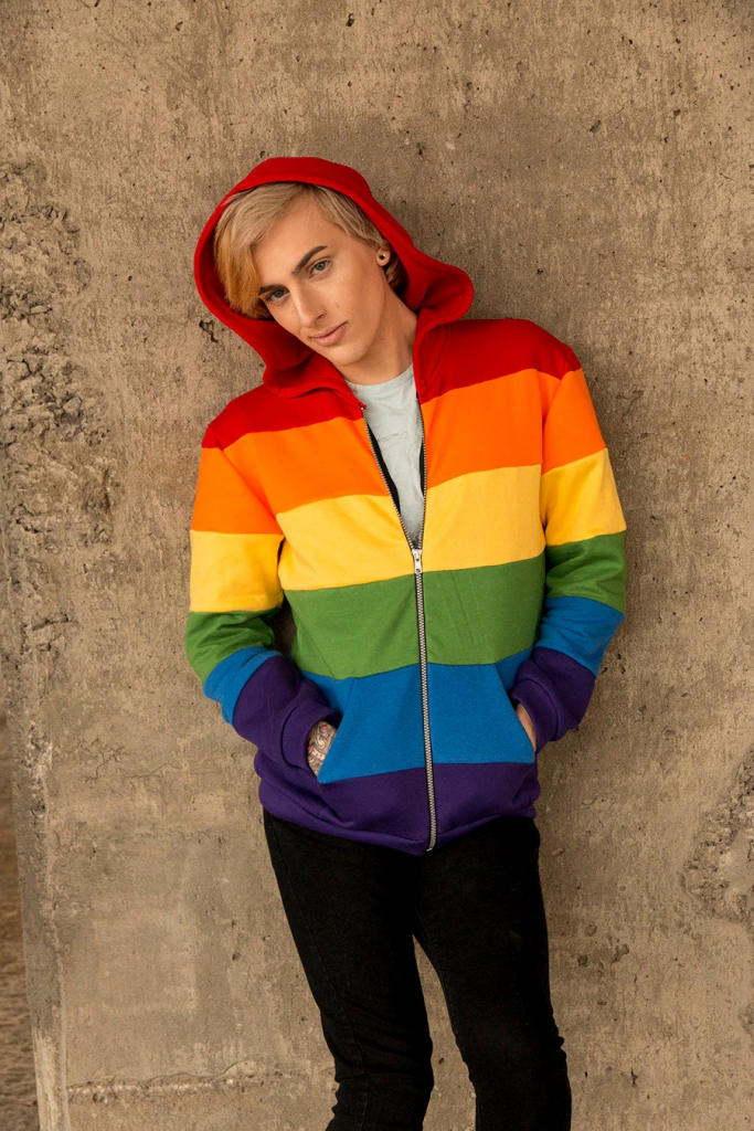 A blond, white person standing in front of a wall wearing a rainbow zip-up sweatshirt with the hood up.