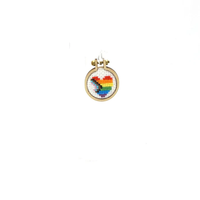A mini embroidery hoop pendant with a progress pride heart in the middle. The flag is in the colors of the rainbow, intersected by a chevron made up of white, light pink, light blue, brown and black stripes. 