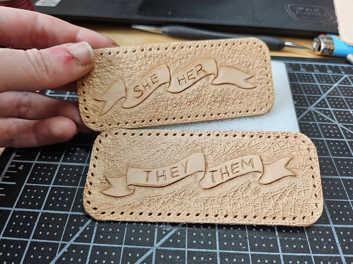 A pair of tan leather patches on a black mat. The top patch, being held up by a hand, is carved with she/her on a banner.
The bottom patch is carved with the/them on a banner. 