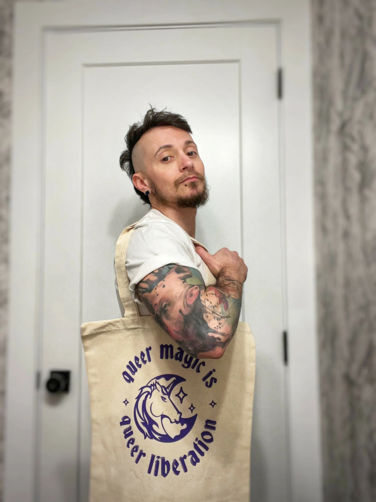A tattooed person standing in front of a door while wearing a tote bag over their shoulder. The tote is tan with purple, small caps lettering which reads, "queer magic is queer liberation." The lettering is curved in a circle around a unicorn crescent moon design.