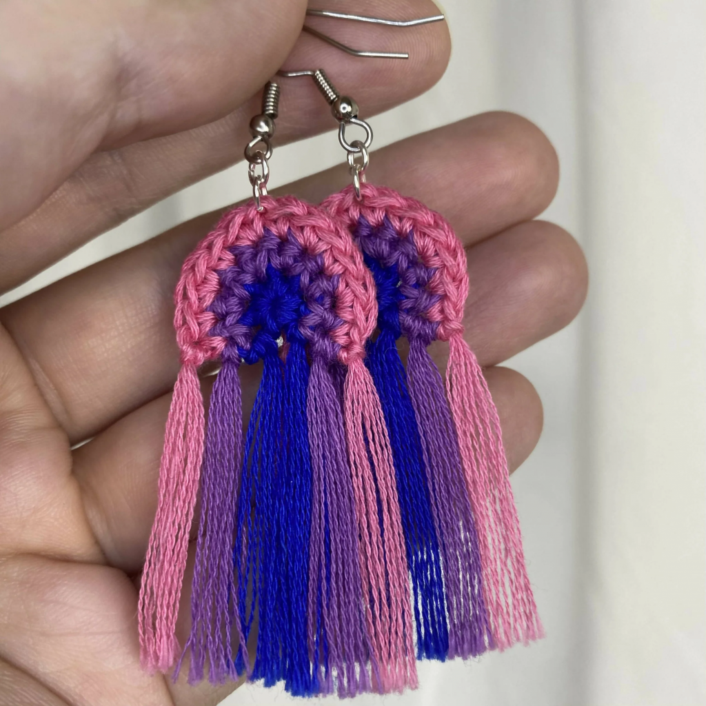 A hand holding up a set of rainbow crochet tassel earrings, with the rainbow made of of the bi flag colors (pink, purple, blue). 